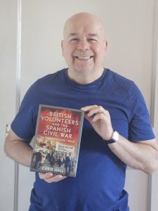 Colour photograph of a white man smiling, holding up a book titled ‘British Volunteers and the Spanish Civil War: ‘The Passionate Cause’, 1936-39’.