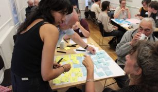 Image of A group of people sit around tables in a bright room. They are collaborating with colourful sticky notes, while two women in the foreground discuss ideas over a shared board.