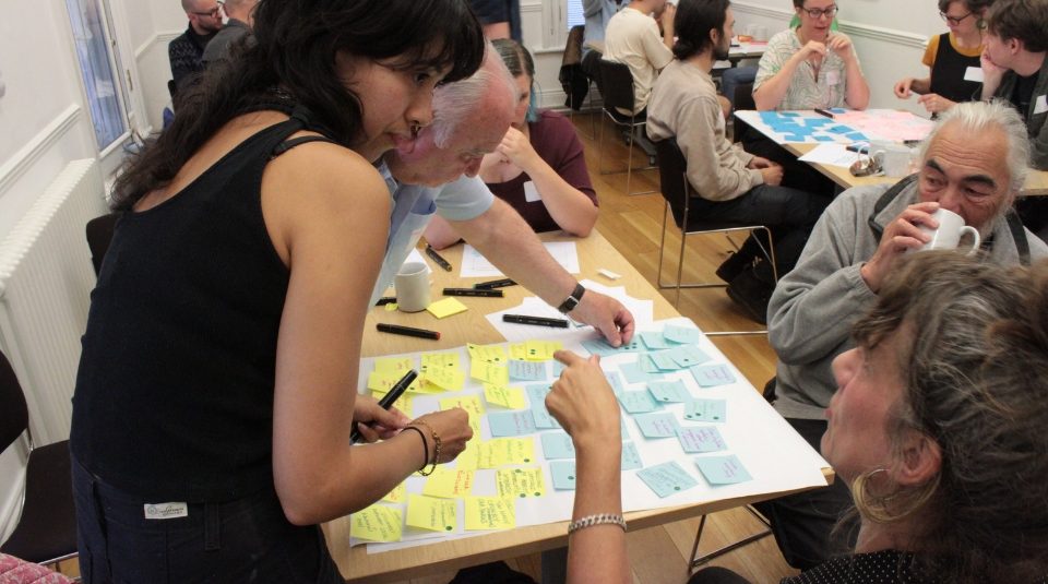 Image of A group of people sit around tables in a bright room. They are collaborating with colourful sticky notes, while two women in the foreground discuss ideas over a shared board.