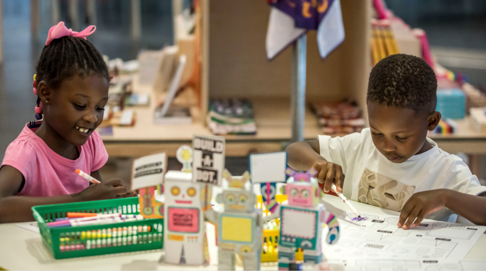Image of Colour photograph of two children colouring in, with paper robot models in the foreground.