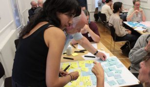 Image of A group of people sit around tables in a bright room. They are collaborating with colourful sticky notes, while two women in the foreground discuss ideas over a shared board.