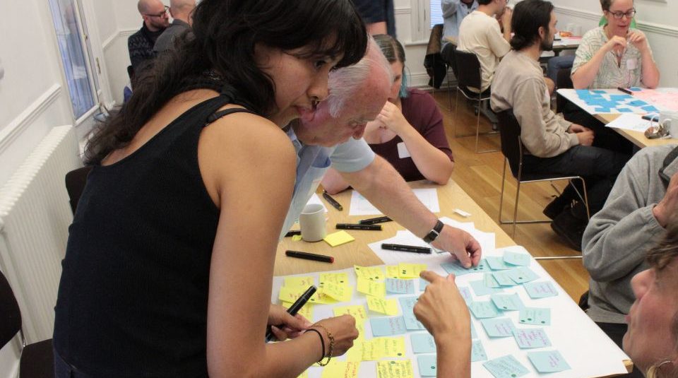Image of A group of people sit around tables in a bright room. They are collaborating with colourful sticky notes, while two women in the foreground discuss ideas over a shared board.