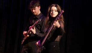 Image of Photograph of two musicians playing electric guitars on stage with dark curtains in background. Both wear black clothing, with one musician playing a bass guitar and the other playing a standard electric guitar.