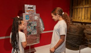 Image of Photograph of two children in a gallery, interacting with a homemade robot model made from cardboard and covered in aluminum foil, decorated with colourful stickers and buttons.