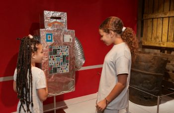 Image of Photograph of two children in a gallery, interacting with a homemade robot model made from cardboard and covered in aluminum foil, decorated with colourful stickers and buttons.