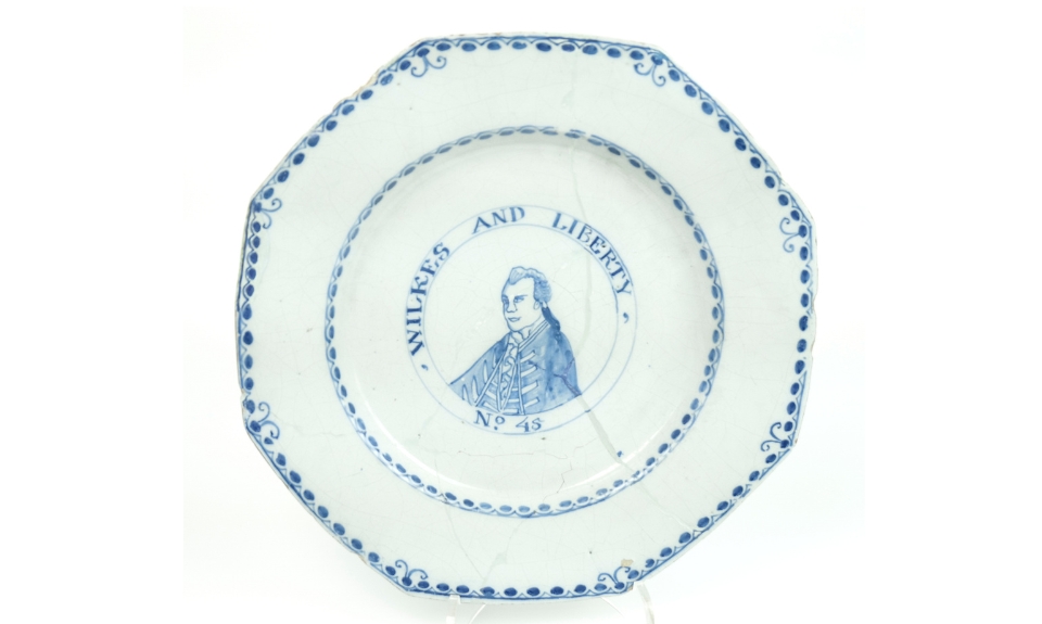 Colour photograph of an octagonal ceramic plate featuring a blue portrait of a man in the centre with the text: 'WILKES AND LIBERTY No 45' surrounding it. The plate has a decorative blue border with small floral patterns and visible cracks on its surface.