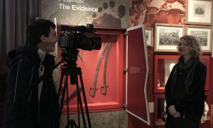 Photograph showing a person filming another individual in a museum setting with historical artifacts displayed in a red case behind them. The display includes two curved swords and several framed pictures on a wall with text partially visible reading Peterloo: The Evidence.