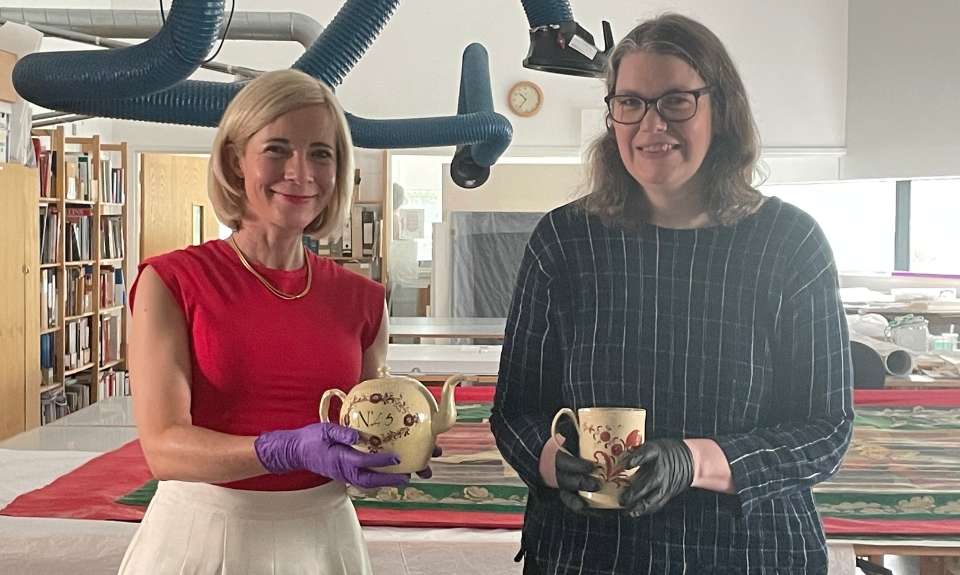 Image of Colour photograph of two people standing in a conservation workspace, each holding a piece of pottery. The setting includes worktables, ventilation hoses, and shelves with books.