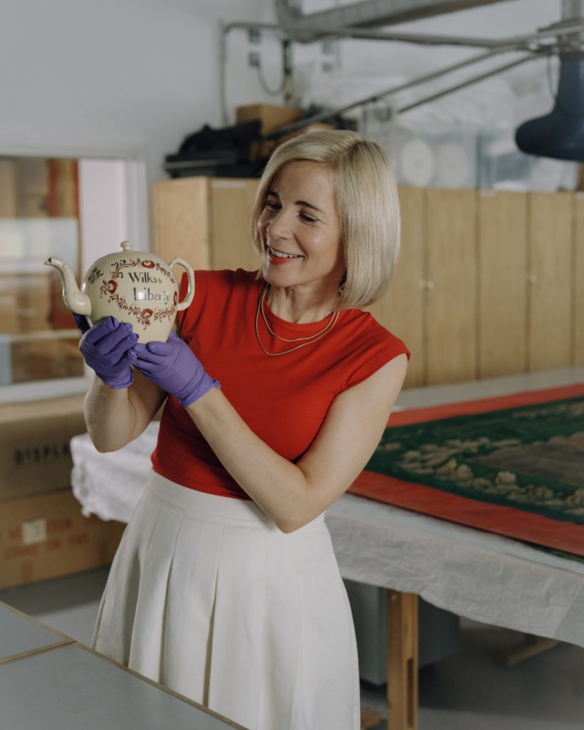 Colour photograph of a person wearing purple gloves and holding a decorative teapot with floral designs and text, in a conservation studio.