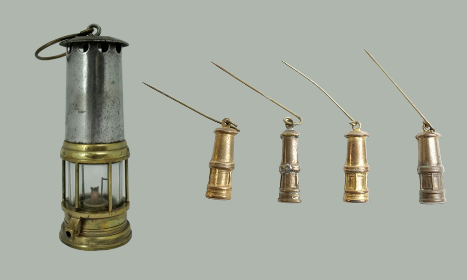 Replica miner's lamp, 1926, and miner's lamp badges, 1926, NMLH 1992 626. Images courtesy of People's History Museum. Colour photograph of a brass miner's lamp, next to four brass badges, each the shape of a miner's lamp.