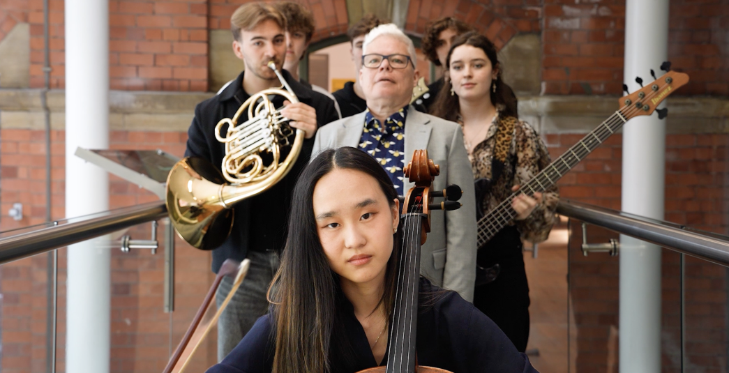 Image of Photograph of a group of five musicians standing indoors against a brick wall, each holding different instruments including a cello, French horn, and bass guitar.
