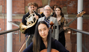 Image of Photograph of a group of five musicians standing indoors against a brick wall, each holding different instruments including a cello, French horn, and bass guitar.