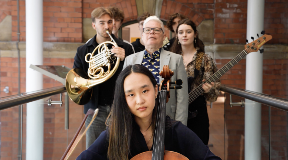 Image of Photograph of a group of five musicians standing indoors against a brick wall, each holding different instruments including a cello, French horn, and bass guitar.