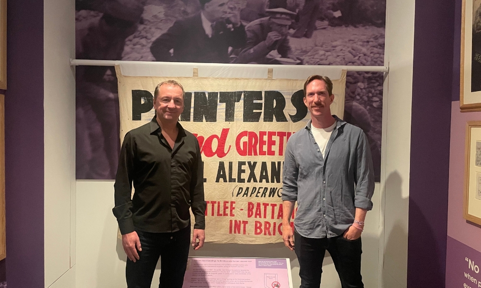 Gideon Long (left) and Ken Appledorn (right) filming at People's History Museum for Spanish TV, 2026. Colour photograph of two people standing in front of a banner on display in a museum gallery.