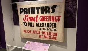 Image of hotograph of a cream banner with bold black and red text reading 'PRINTERS Send GREETINGS TO BILL ALEXANDER (PAPERWORKERS) MAJOR ATTLEE - BATTALION INT. BRIGADE.'