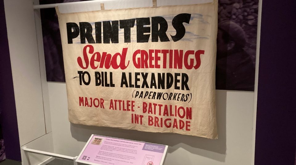 Image of hotograph of a cream banner with bold black and red text reading 'PRINTERS Send GREETINGS TO BILL ALEXANDER (PAPERWORKERS) MAJOR ATTLEE - BATTALION INT. BRIGADE.'