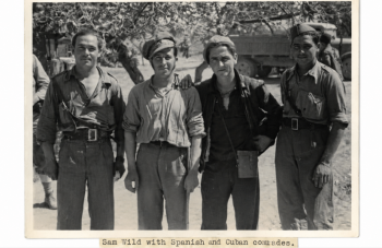 Image of Black and white photograph of four standing men wearing army uniform, with a tree and a truck in the background.