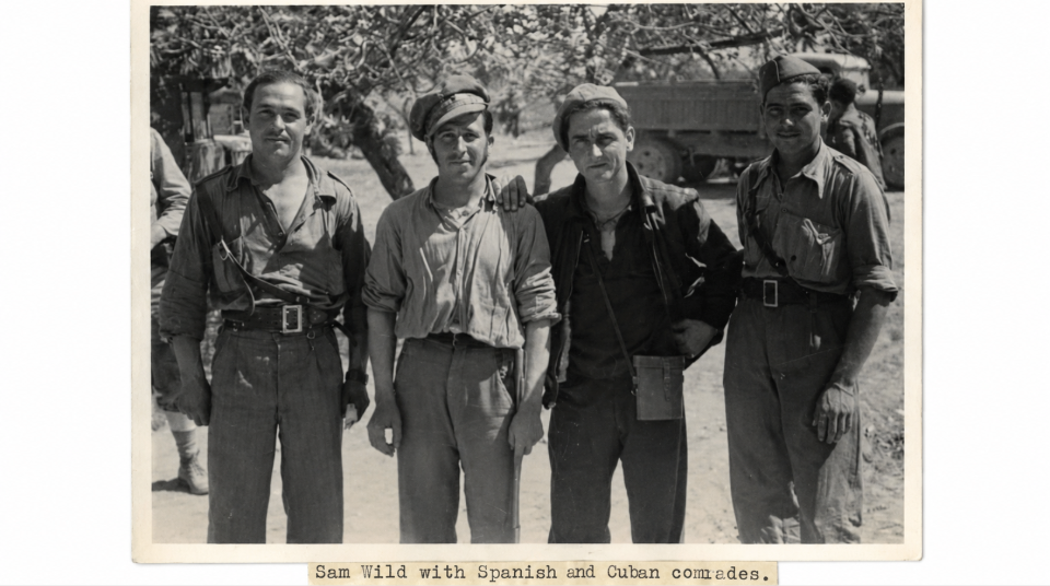 Image of Black and white photograph of four standing men wearing army uniform, with a tree and a truck in the background.