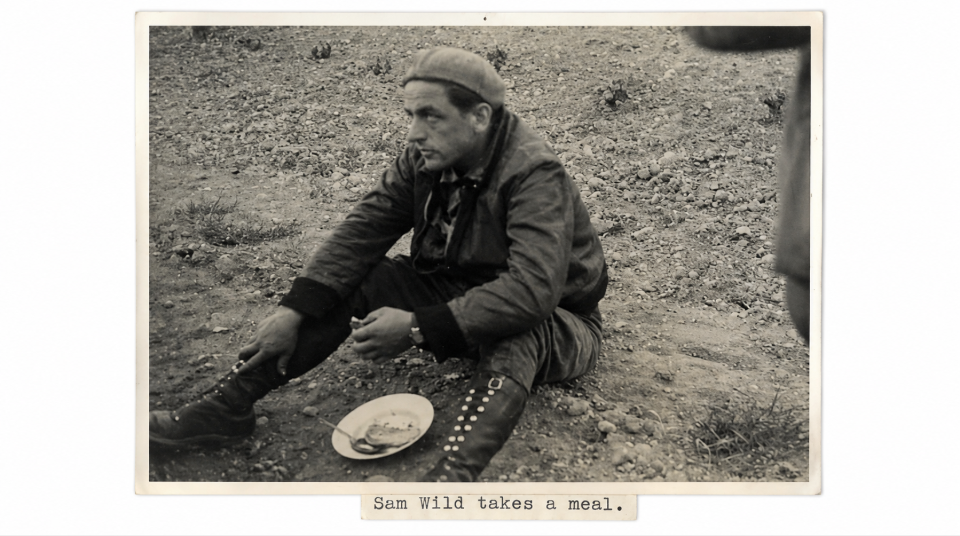 Sam Wild in Spain, around 1937. Image courtesy of People's History Museum. Black and white photograph of a man wearing army uniform sitting on the ground eating food from a plate, with a typed caption reading: 'Sam Wild takes a meal.'.