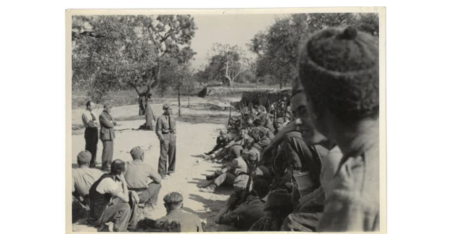 Sam Wild, Bob Cooney, Bill Rust and others, Spain, around 1937. Image courtesy of People's History Museum. Black and white photograph including a man stood up talking to a group of seated men - all wearing army uniform.