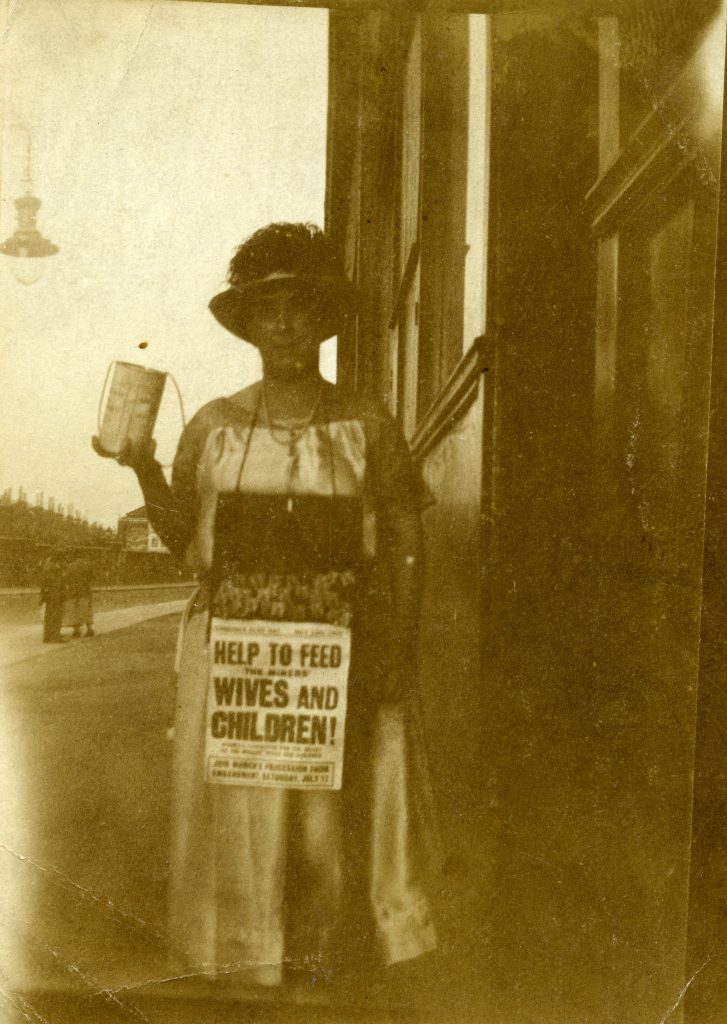 Women’s Committee for the Relief of Miners’ Wives and Children fundraising photograph, 1926. Image courtesy of People's History Museum. Black and white photograph of a woman holding a tin to collect funds, wearing a sign including text: 'Help To Feed The Miners' Wives And Children!'