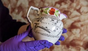 Image of A ceramic jug featuring a printed map design with detailed street names and buildings, including 'Peter Street'. The jug has a floral pattern around the rim and is held by hands wearing purple gloves.