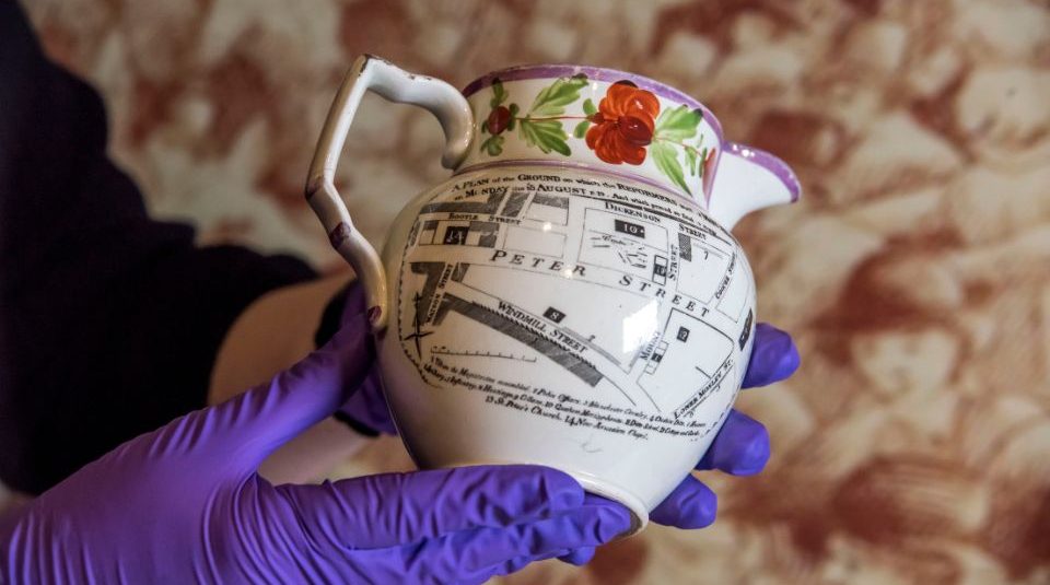 Image of A ceramic jug featuring a printed map design with detailed street names and buildings, including 'Peter Street'. The jug has a floral pattern around the rim and is held by hands wearing purple gloves.