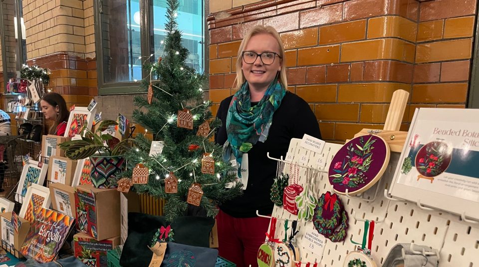 Image of A stallholder standing in front of a display of textile gifts she is selling at the market.