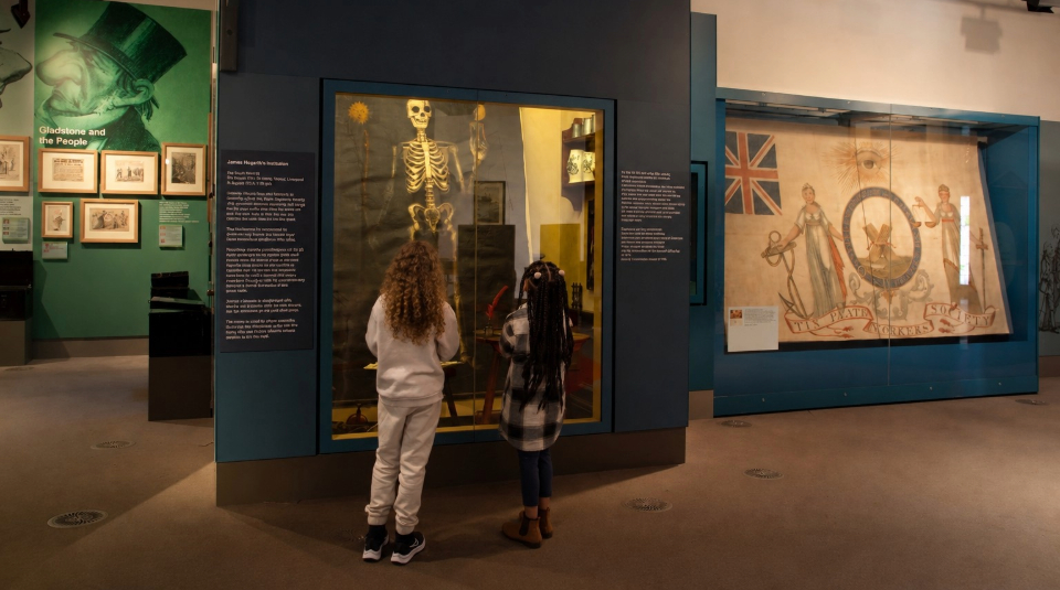 Image of Two young visitors exploring a display including a skeleton painting in a museum gallery.