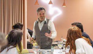 Image of Colour photograph of a man holding a glass bottle, standing and talking to a table of seated people.