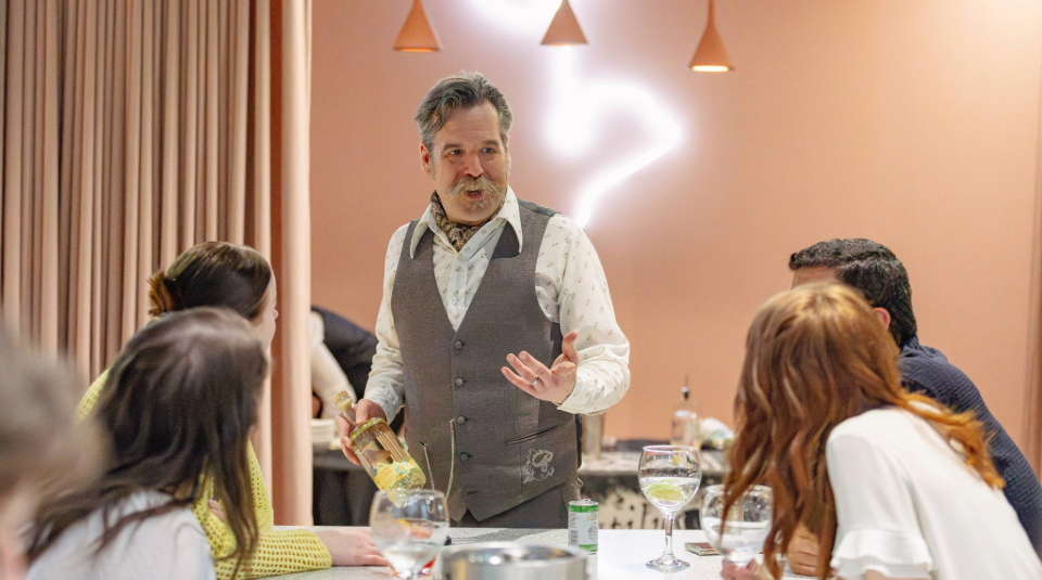 Image of Colour photograph of a man holding a glass bottle, standing and talking to a table of seated people.