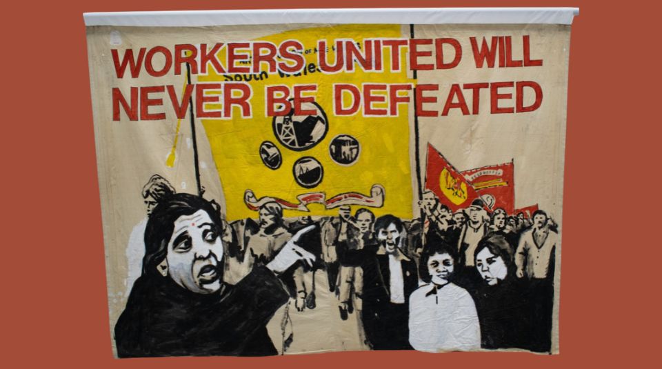 Image of A painted banner, featuring the slogan ‘WORKERS UNITED WILL NEVER BE DEFEATED’ and an illustration of a crowd protesting.