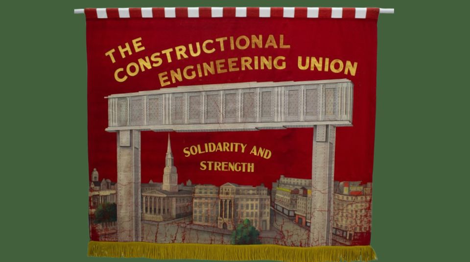 Image of A red banner with gold lettering reading: ‘THE CONSTRUCTIONAL ENGINEERING UNION’ and ‘SOLIDARITY AND STRENGTH’, with an illustration of a large bridge like structure supported by pillars, with a cityscape below.